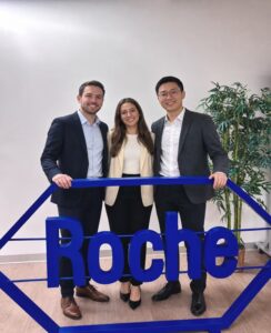 Three professionals pose together behind a blue Roche sign in a bright office.